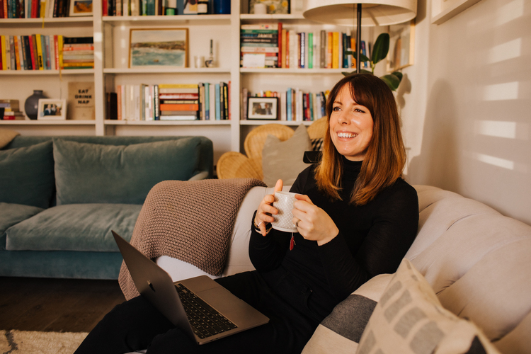 Amy sits on her sofa smiling and holding a cup of tea. Her laptop is open on her lap. a colourful bookshelf is visible in the background