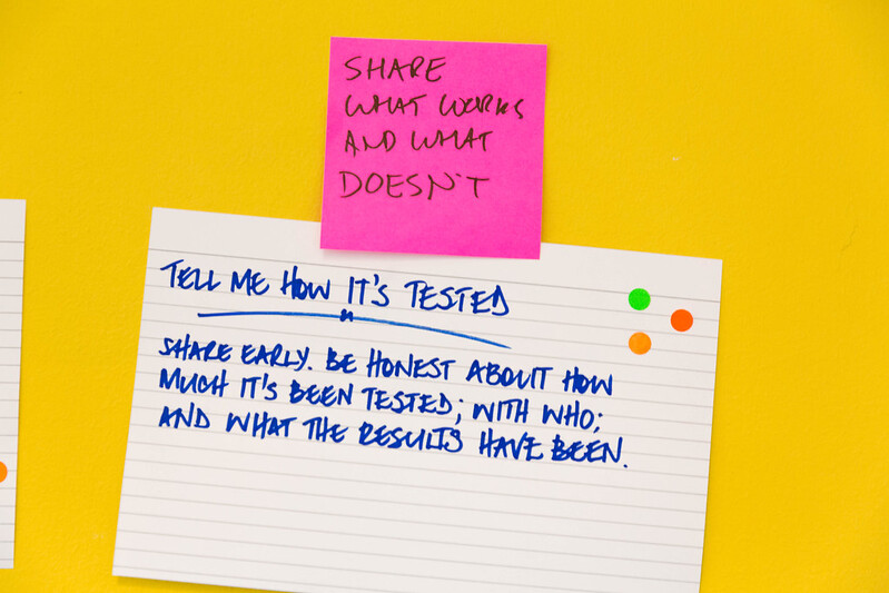 a pink post-it on a yellow background. The post-is reads 'share what works and what doesn't' and underneath it is a hand-written note that says 'tell me how it's been tested. Share early, be honest about how much it's been tested, with who, and what the results has been.'