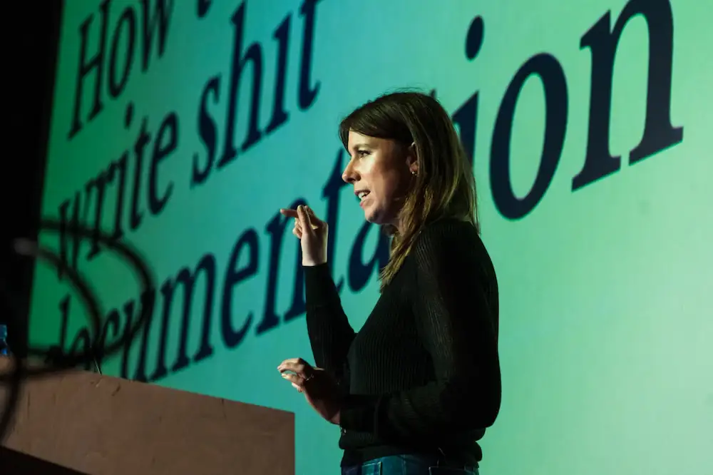 Amy delivering a talk in front of a large screen. The text on the screen says How To Write Shit Documentation.
